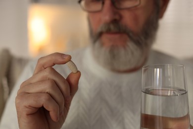 Photo of Senior man with pill and glass of water indoors, selective focus