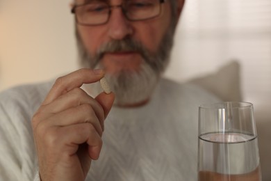 Photo of Senior man with pill and glass of water indoors, selective focus