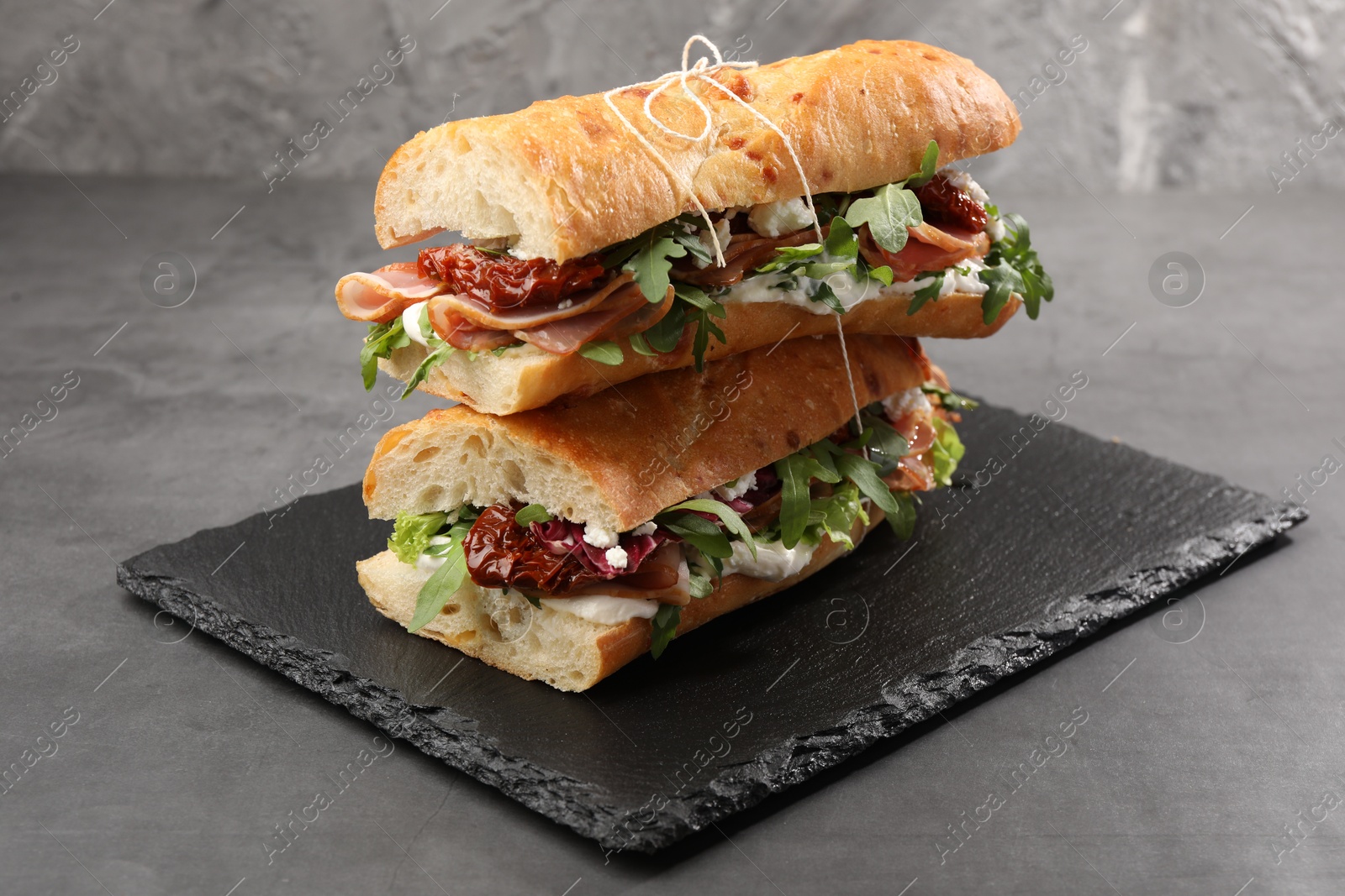 Photo of Halves of baguette sandwich with prosciutto and sun dried tomatoes on grey table