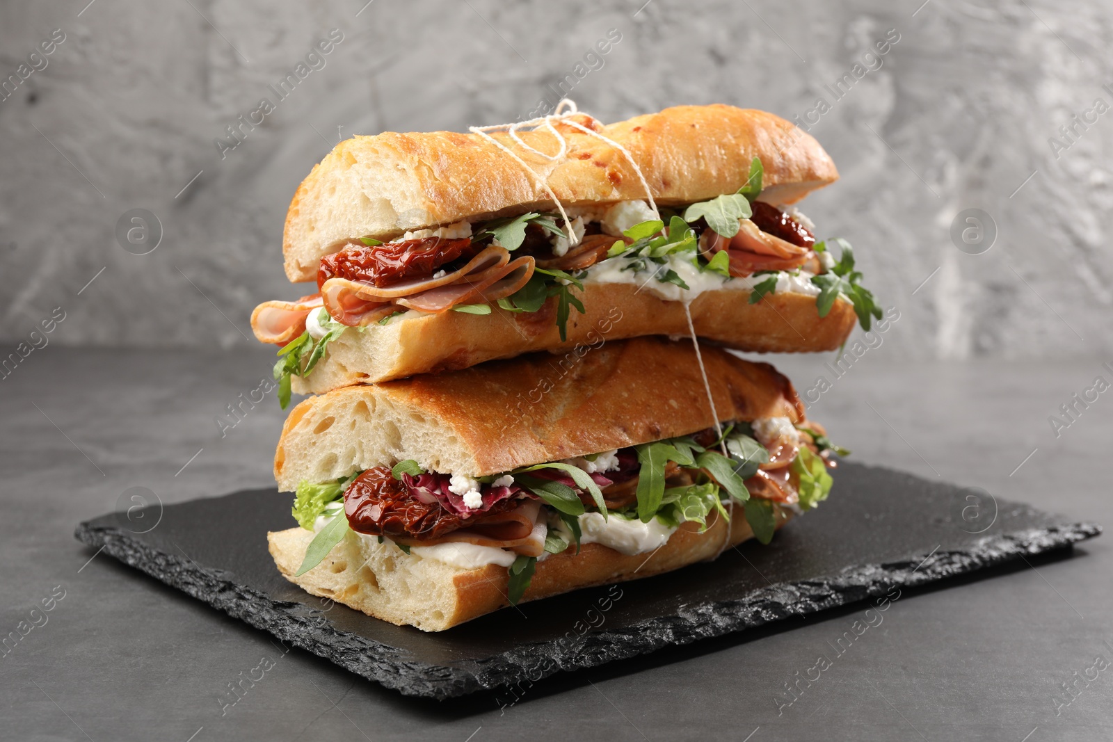 Photo of Halves of baguette sandwich with prosciutto and sun dried tomatoes on grey table