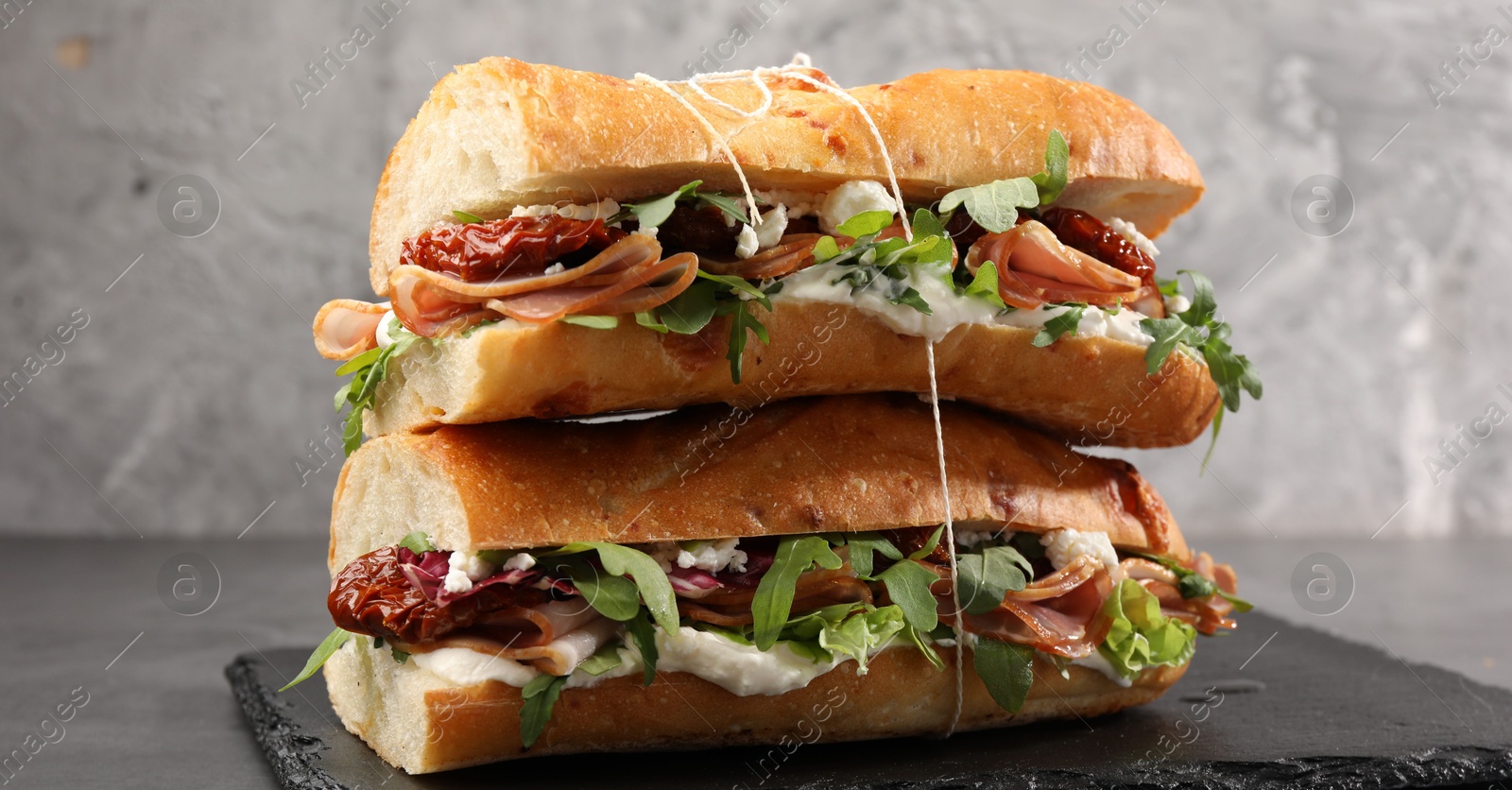 Photo of Halves of baguette sandwich with prosciutto and sun dried tomatoes on grey table, closeup