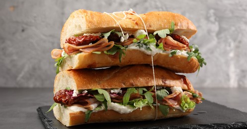 Photo of Halves of baguette sandwich with prosciutto and sun dried tomatoes on grey table, closeup