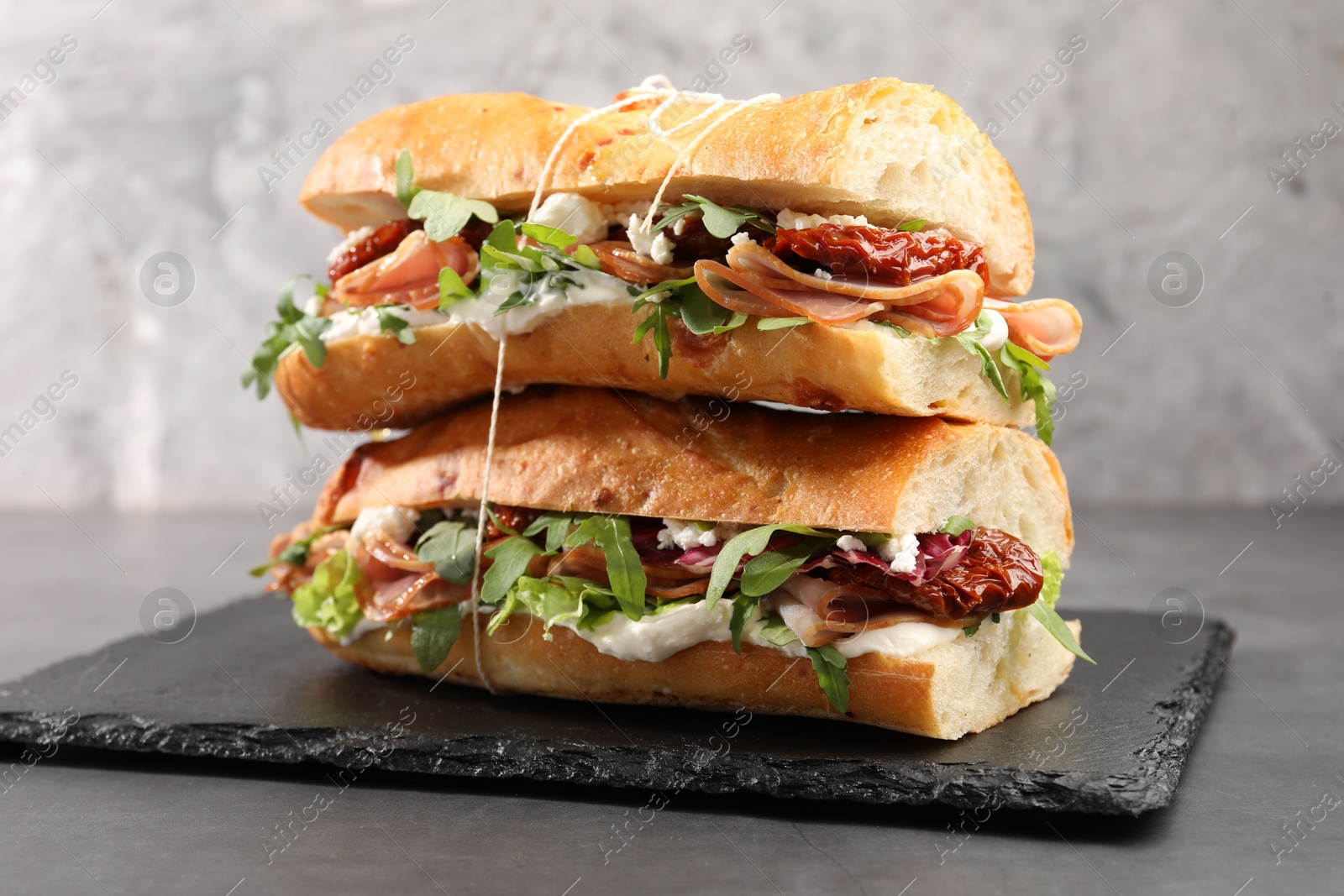 Photo of Halves of baguette sandwich with prosciutto and sun dried tomatoes on grey table, closeup