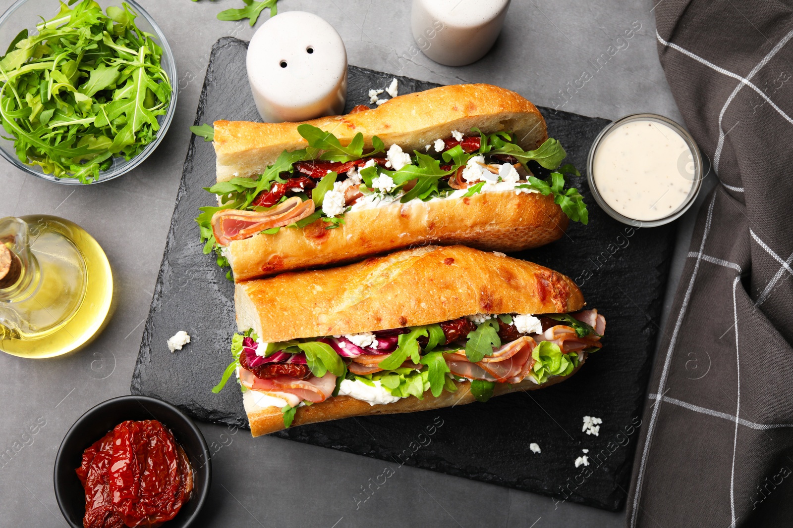 Photo of Halves of baguette sandwich with prosciutto and sun dried tomatoes and ingredients on grey table, flat lay