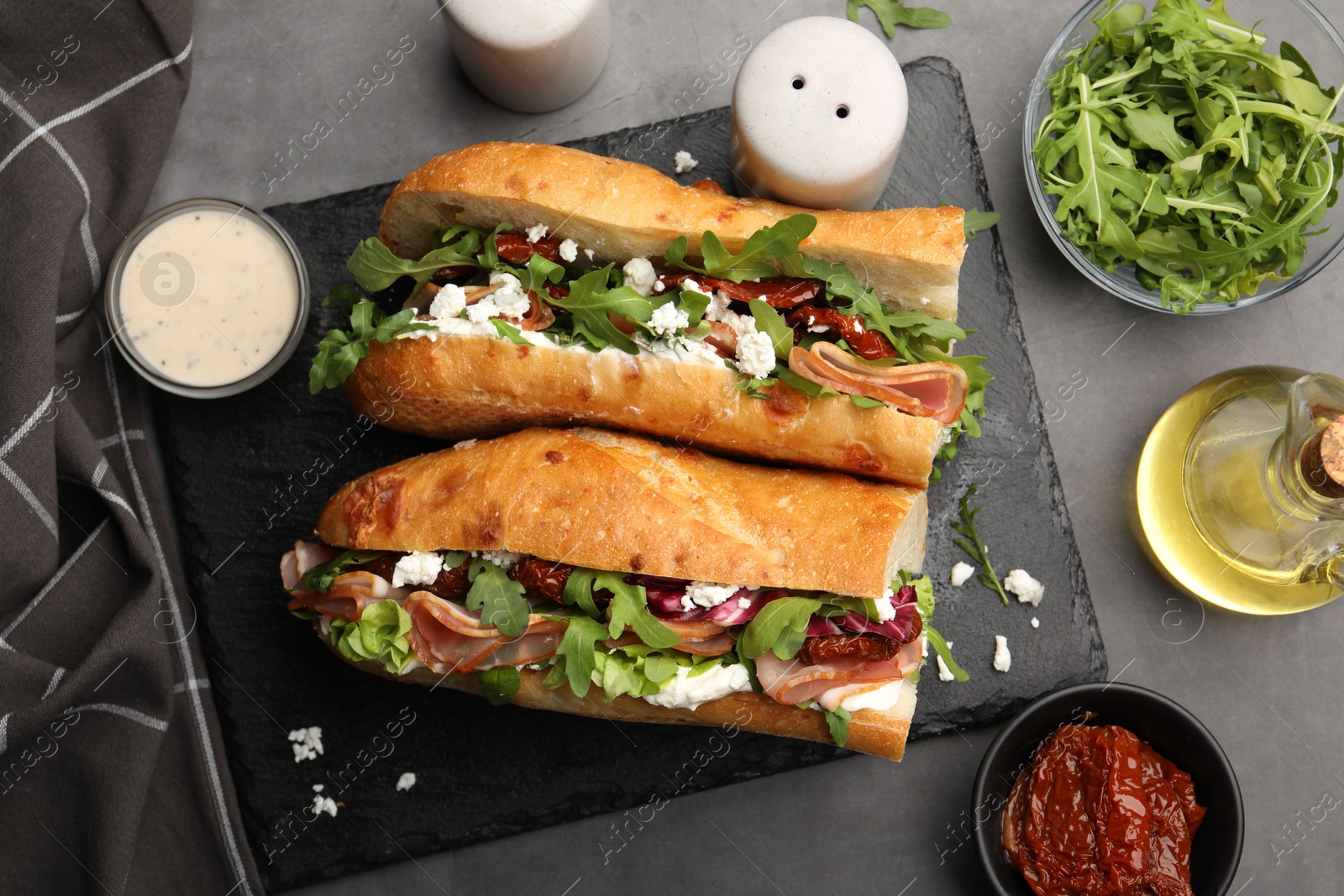 Photo of Halves of baguette sandwich with prosciutto and sun dried tomatoes and ingredients on grey table, flat lay