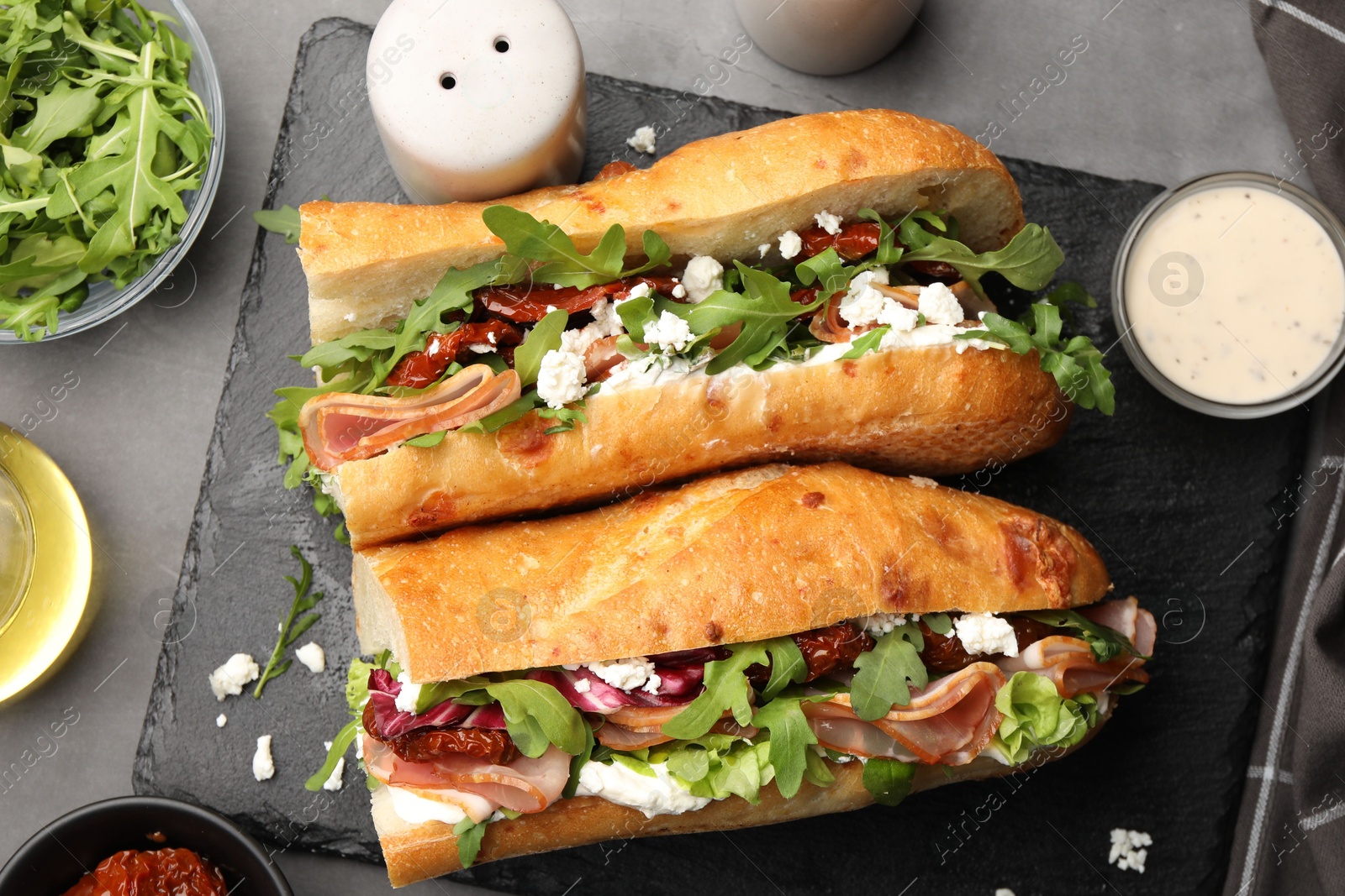 Photo of Halves of baguette sandwich with prosciutto and sun dried tomatoes and ingredients on grey table, flat lay