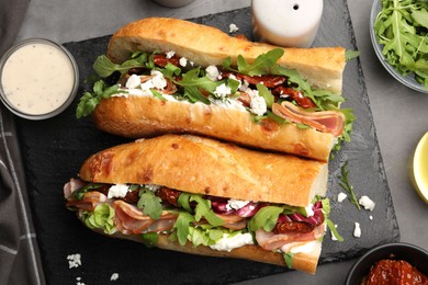 Photo of Halves of baguette sandwich with prosciutto and sun dried tomatoes and ingredients on grey table, flat lay