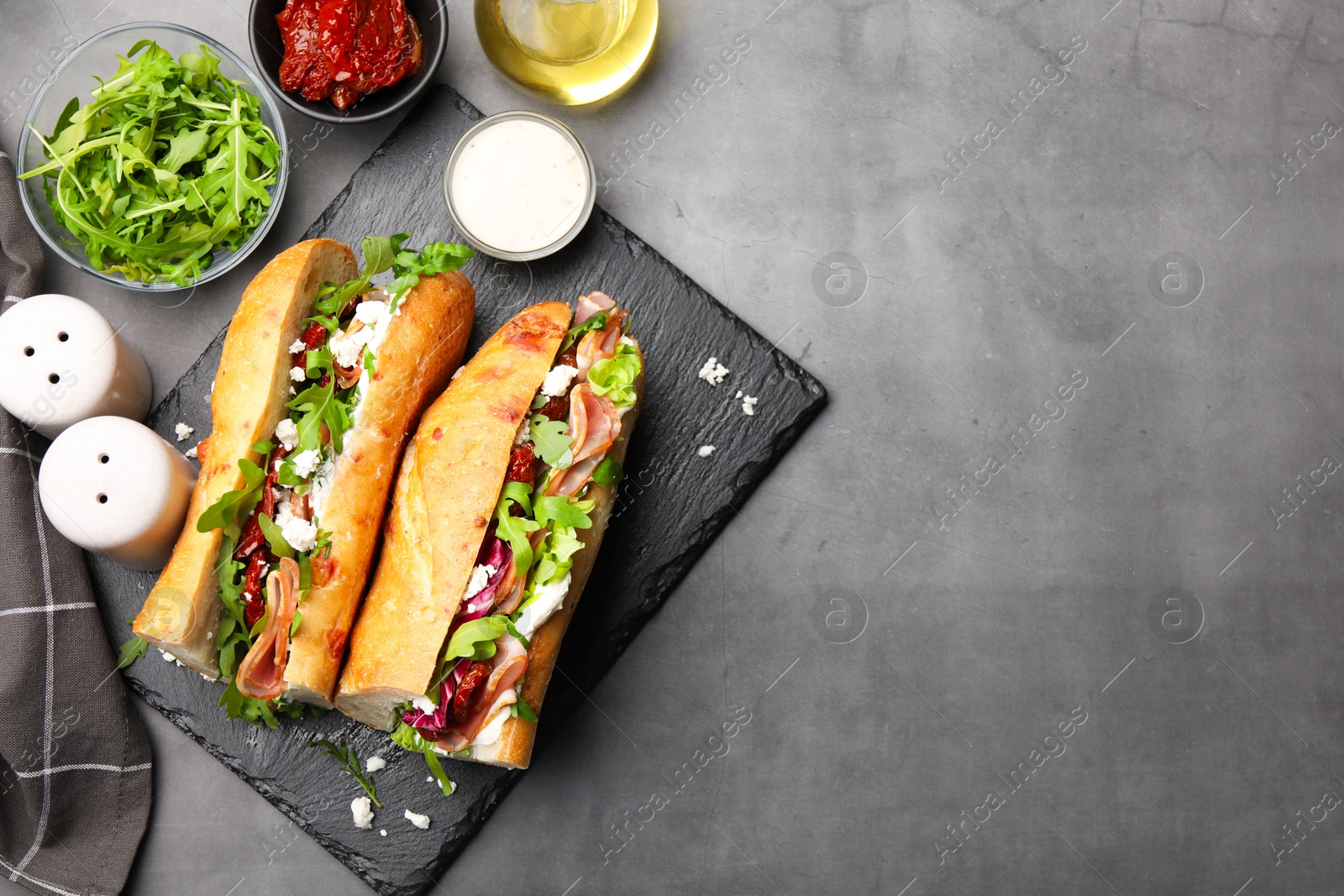 Photo of Halves of baguette sandwich with prosciutto and sun dried tomatoes and ingredients on grey table, flat lay. Space for text