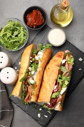 Photo of Halves of baguette sandwich with prosciutto and sun dried tomatoes and ingredients on grey table, flat lay