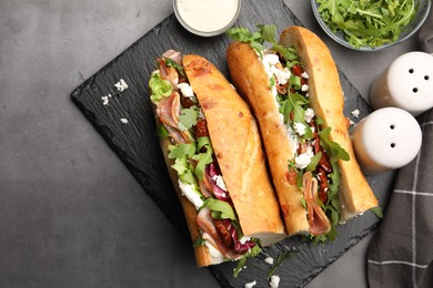 Photo of Halves of baguette sandwich with prosciutto and sun dried tomatoes and ingredients on grey table, flat lay