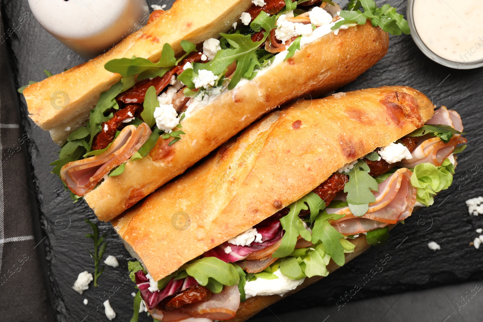 Photo of Halves of baguette sandwich with prosciutto and sun dried tomatoes and ingredients on grey table, flat lay