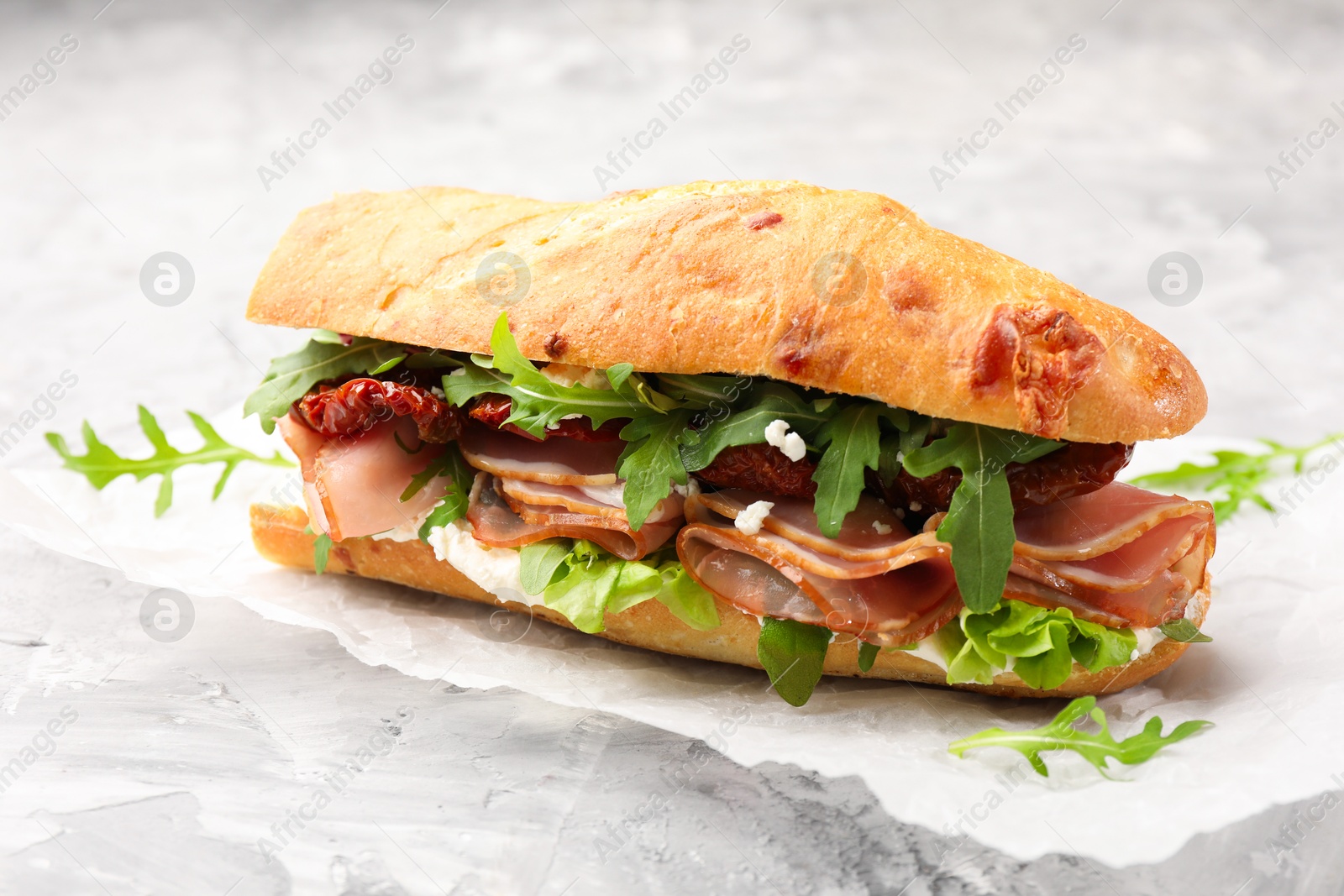 Photo of Baguette sandwich with ham and sun dried tomatoes on grey textured table, closeup