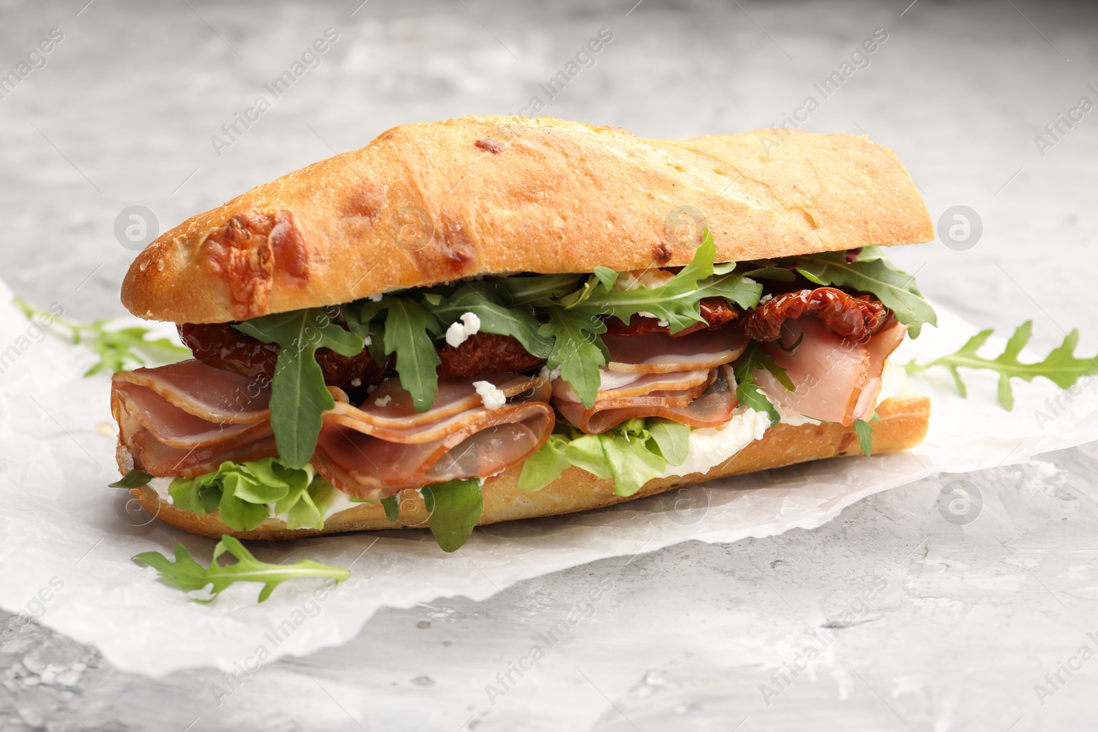 Photo of Baguette sandwich with ham and sun dried tomatoes on grey textured table, closeup