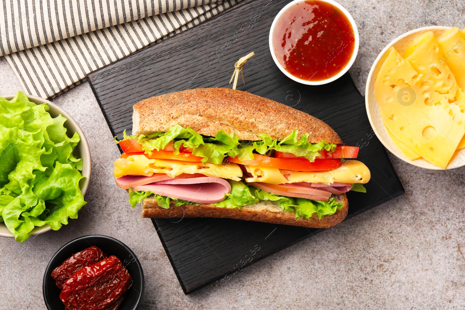 Photo of Delicious baguette sandwich with ham and ingredients on grey table, flat lay