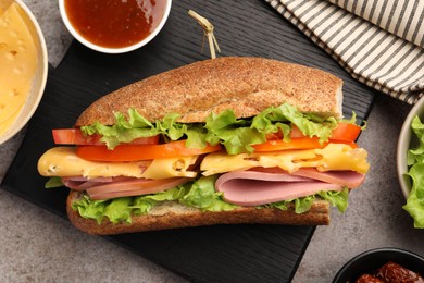 Photo of Delicious baguette sandwich with ham and ingredients on grey table, flat lay