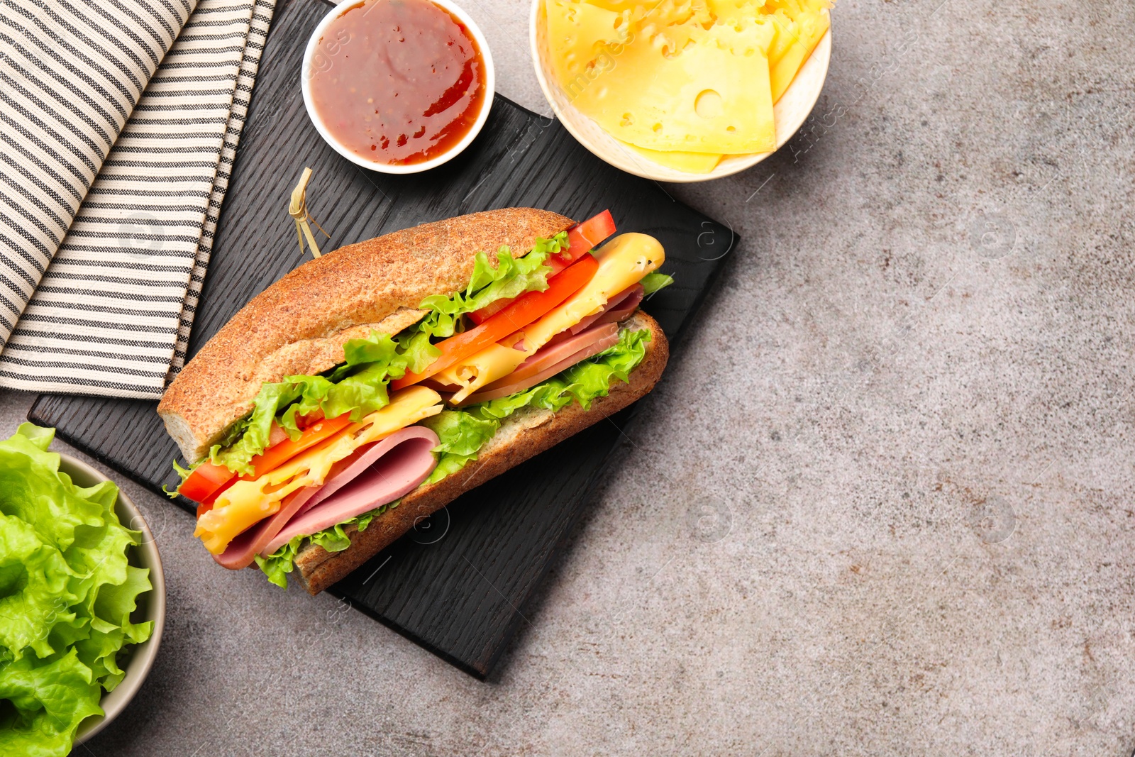 Photo of Delicious baguette sandwich with ham and ingredients on grey table, flat lay. Space for text