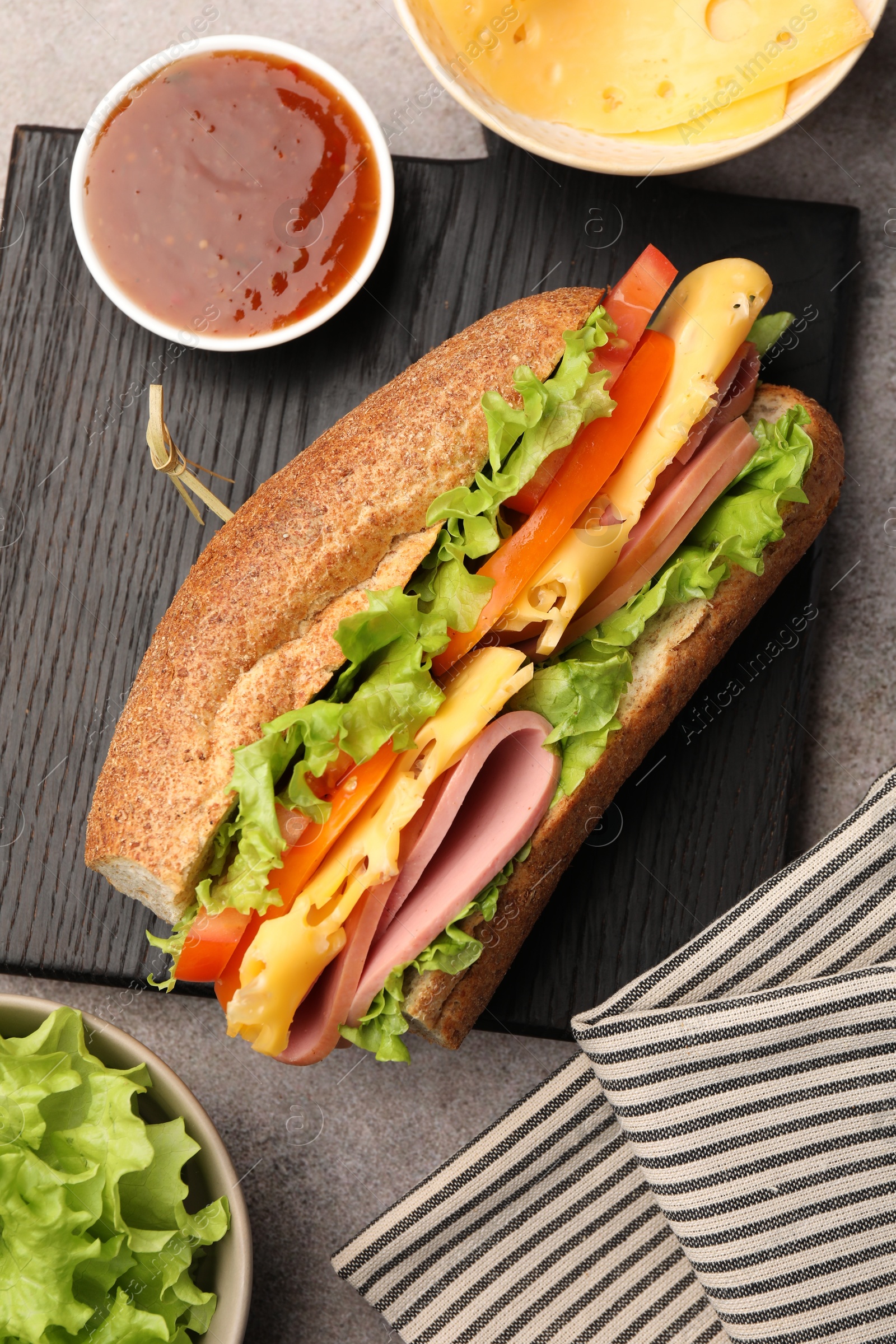 Photo of Delicious baguette sandwich with ham and ingredients on grey table, flat lay