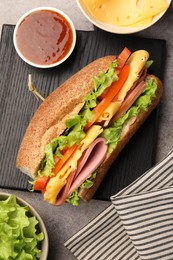 Photo of Delicious baguette sandwich with ham and ingredients on grey table, flat lay