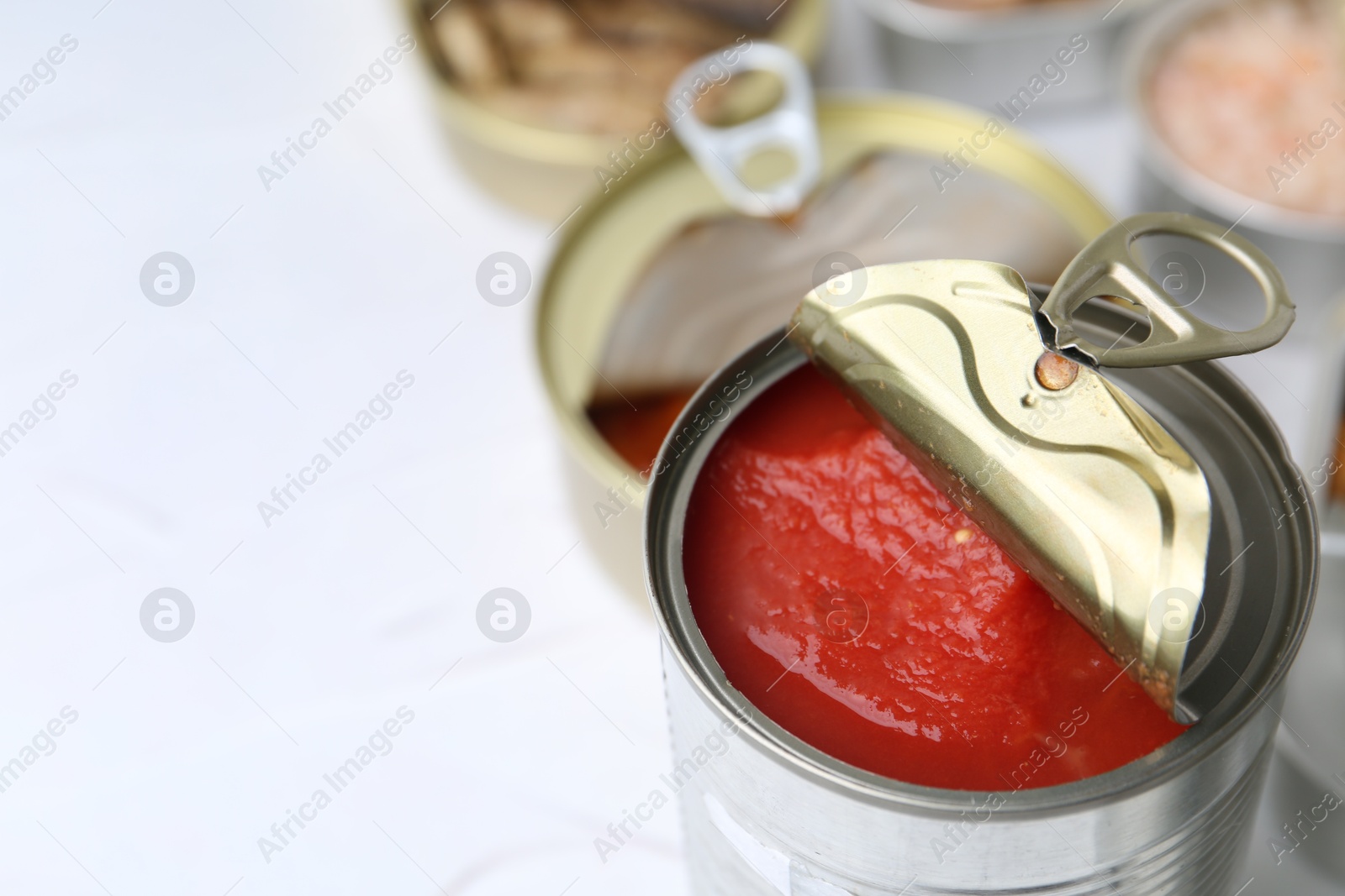 Photo of Open tin cans with different preserved products on white table, closeup. Space for text