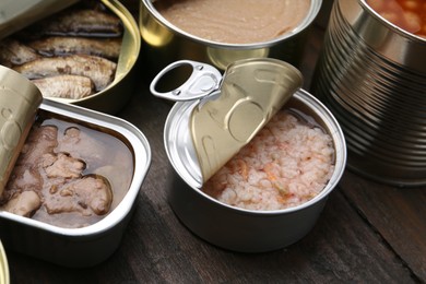 Photo of Open tin cans with different preserved products on wooden table, closeup