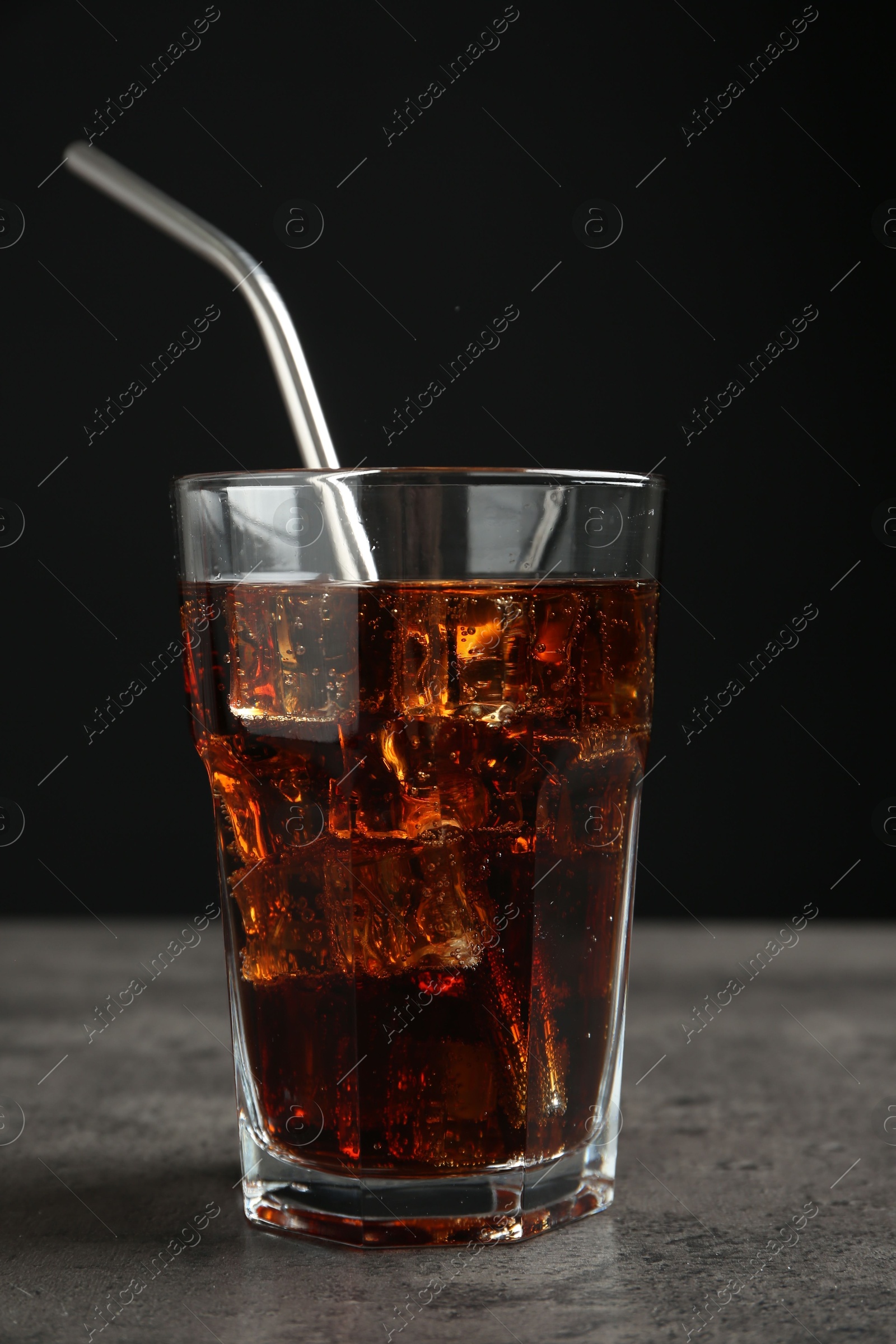 Photo of Cold cola with ice cubes and drinking straw in glass on grey table against black background, closeup