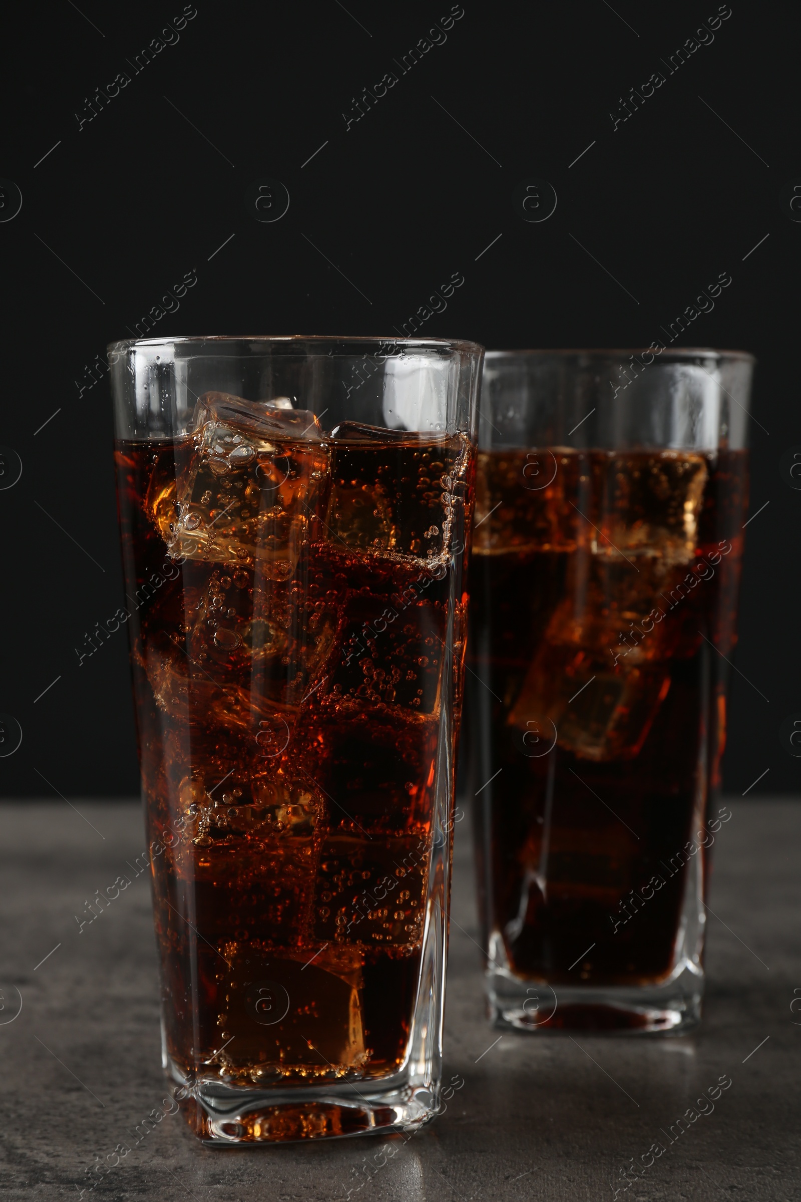 Photo of Cold cola with ice cubes in glasses on grey table against black background, closeup