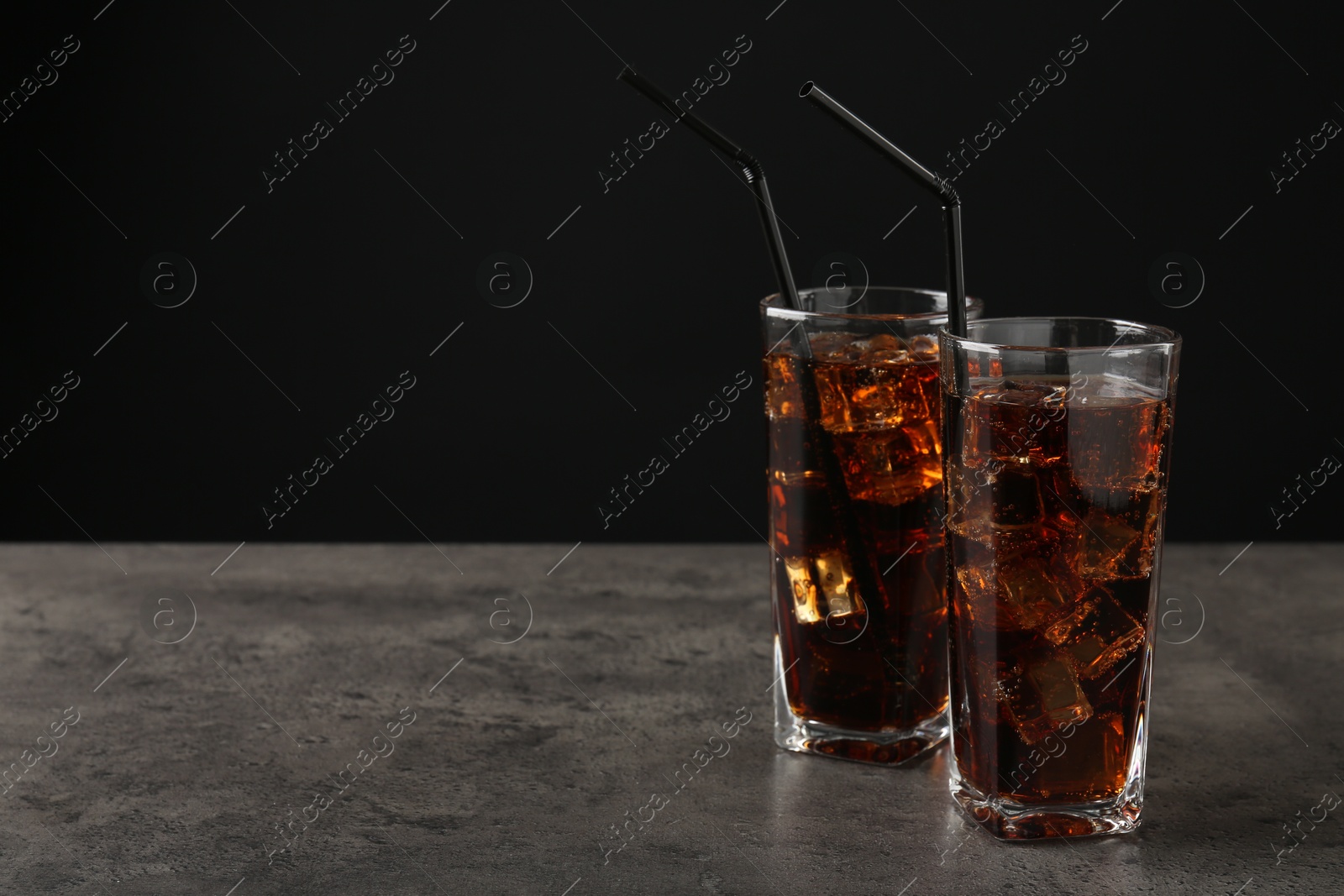 Photo of Cold cola with ice cubes and drinking straws in glasses on grey table against black background. Space for text