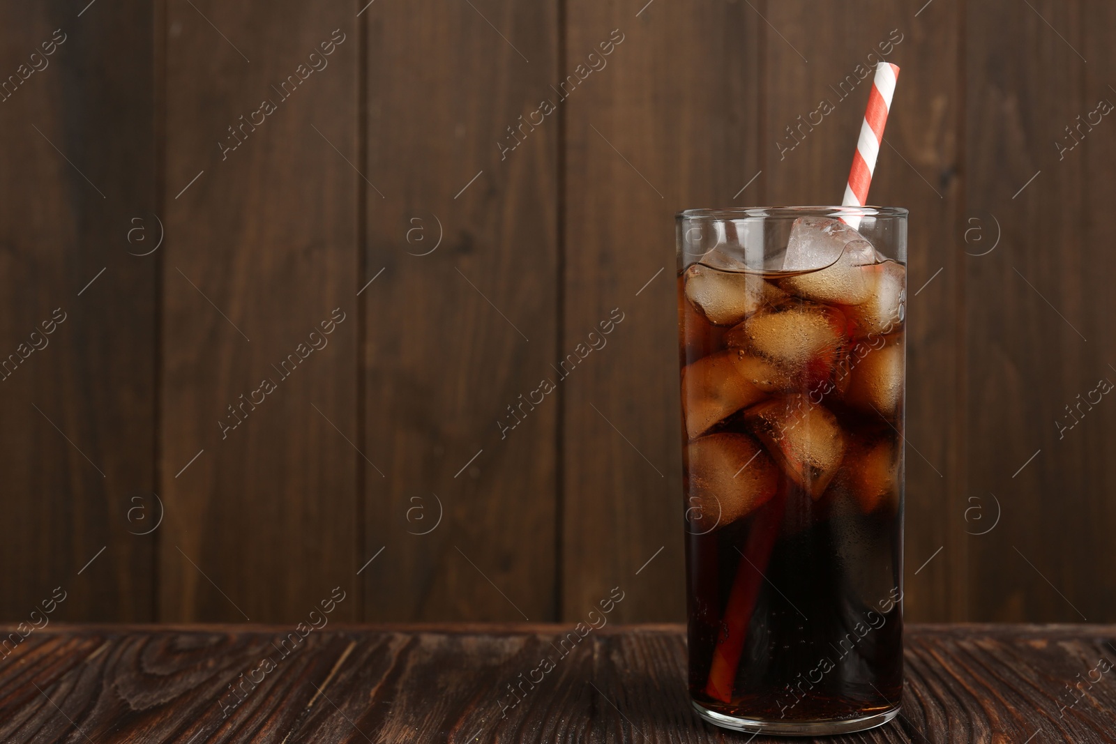 Photo of Cold cola with ice cubes and drinking straw in glass on wooden table. Space for text