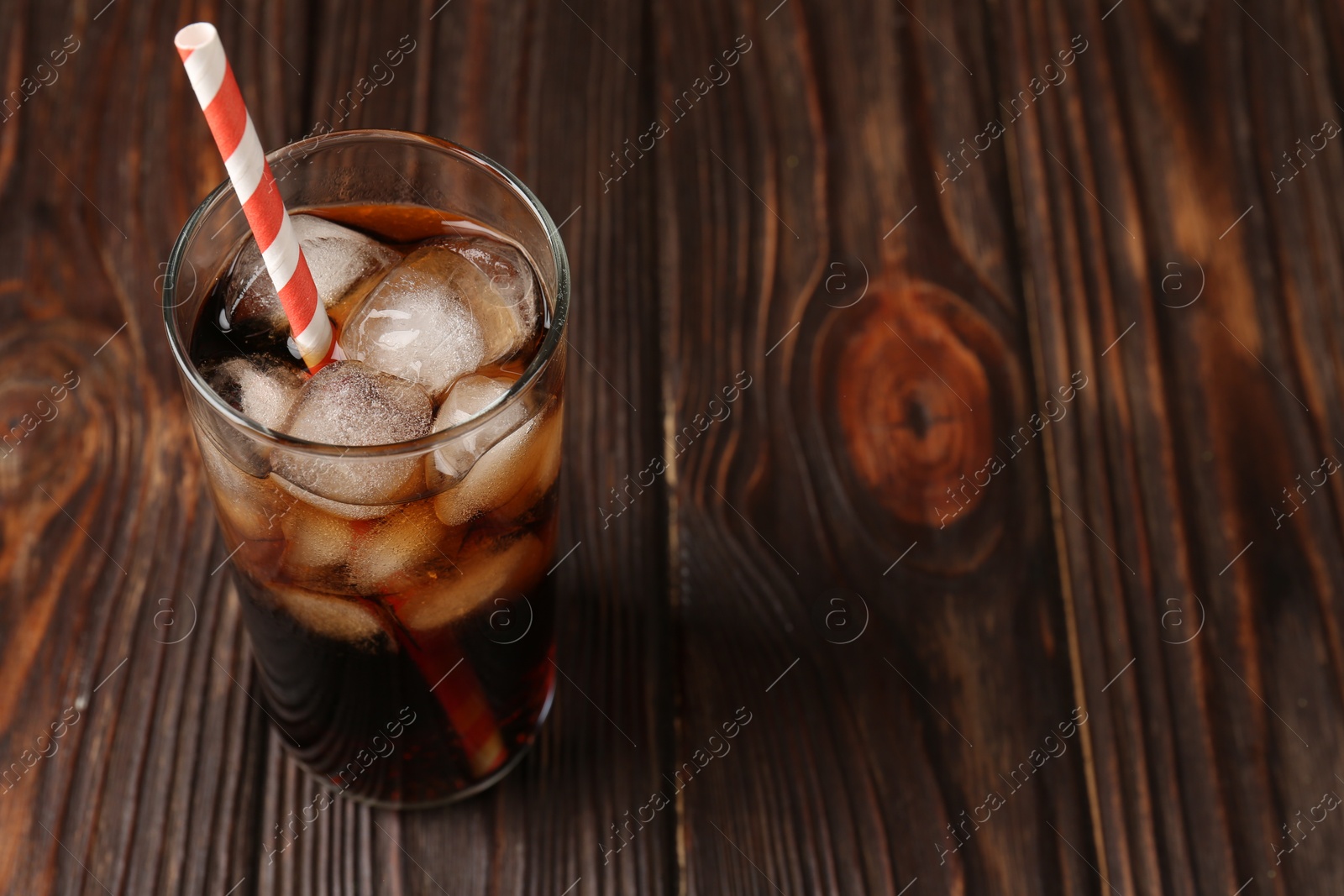 Photo of Cold cola with ice cubes and drinking straw in glass on wooden table. Space for text