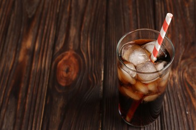 Photo of Cold cola with ice cubes and drinking straw in glass on wooden table. Space for text