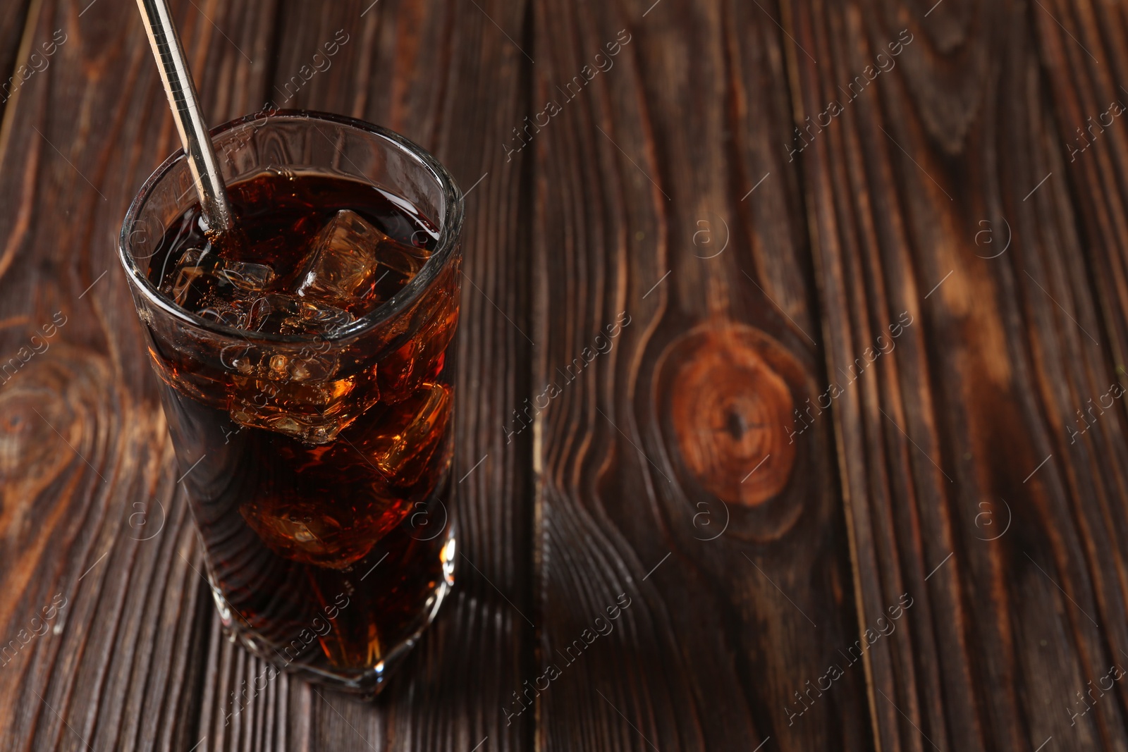 Photo of Cold cola with ice cubes and drinking straw in glass on wooden table. Space for text