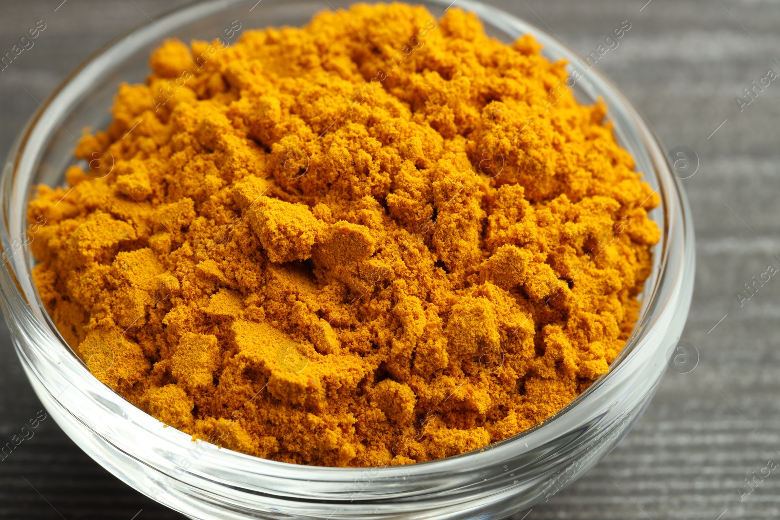 Photo of Turmeric powder in glass bowl on grey wooden table, closeup