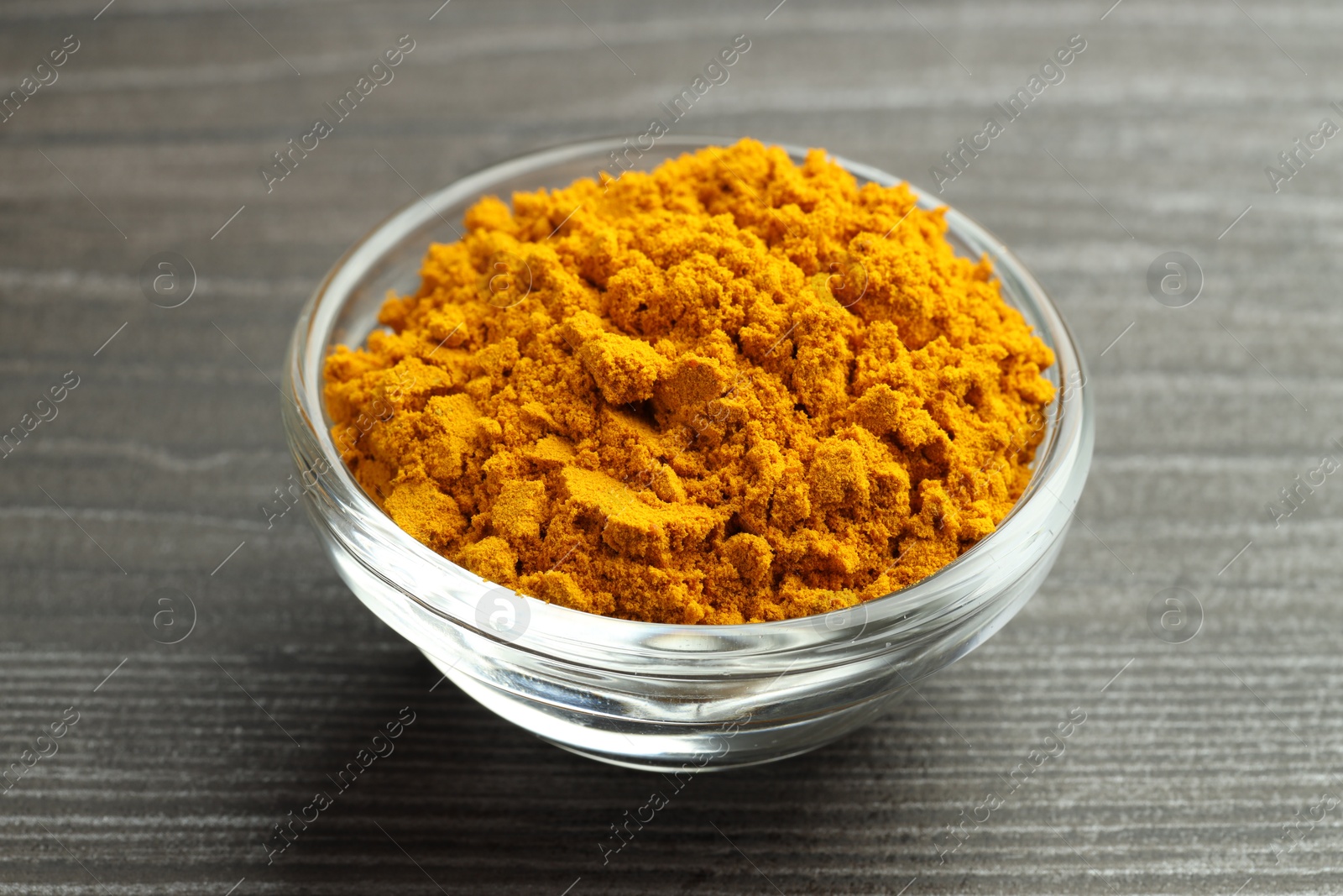 Photo of Turmeric powder in glass bowl on grey wooden table, closeup