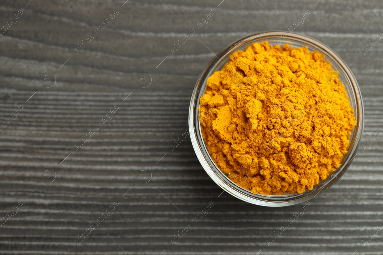 Photo of Turmeric powder in glass bowl on grey wooden table, top view. Space for text