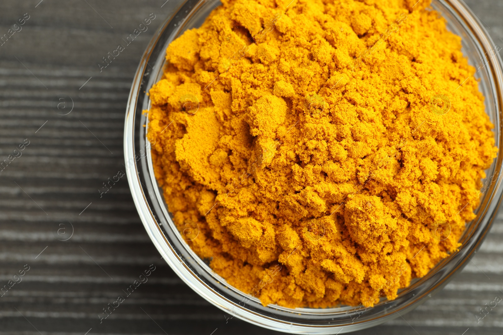 Photo of Turmeric powder in glass bowl on grey wooden table, closeup. Top view