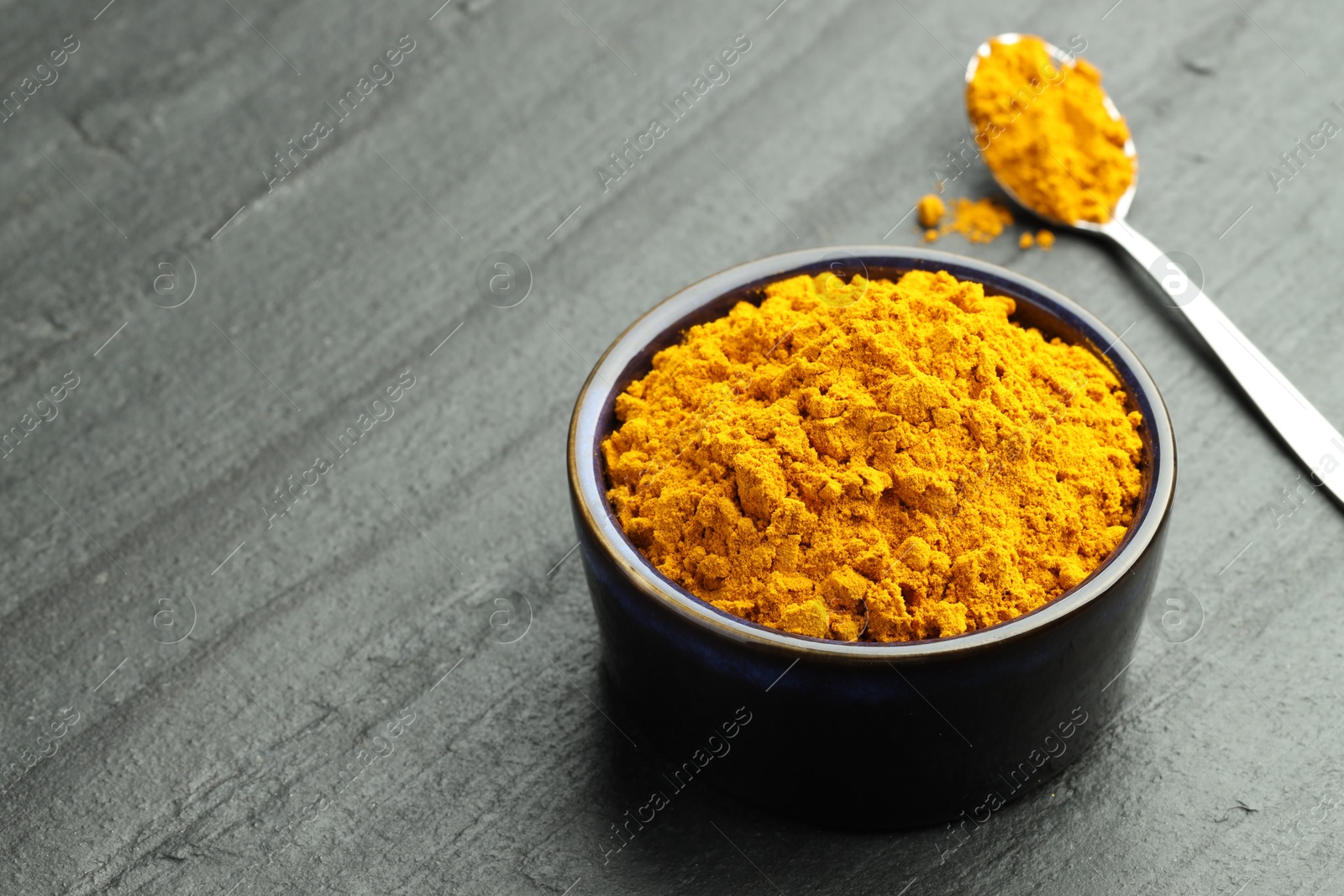 Photo of Turmeric powder in bowl and spoon on grey table, closeup. Space for text