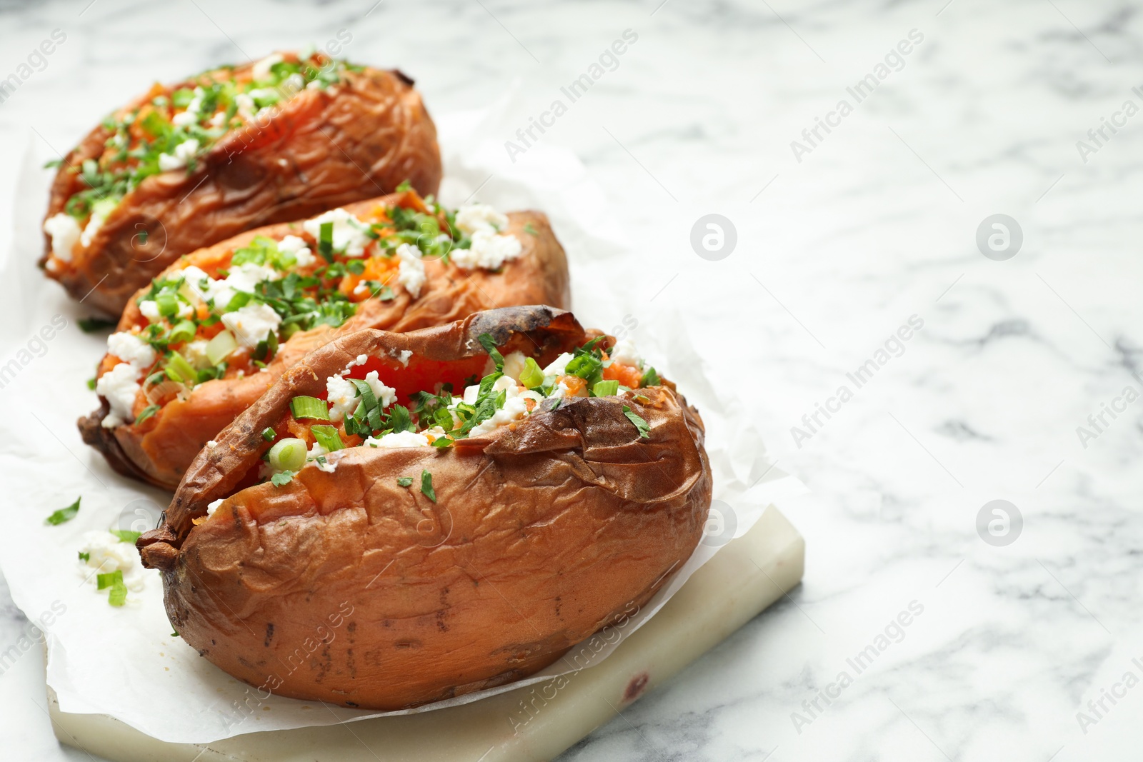 Photo of Tasty cooked sweet potatoes with feta cheese, green onion and parsley on white marble table, closeup. Space for text