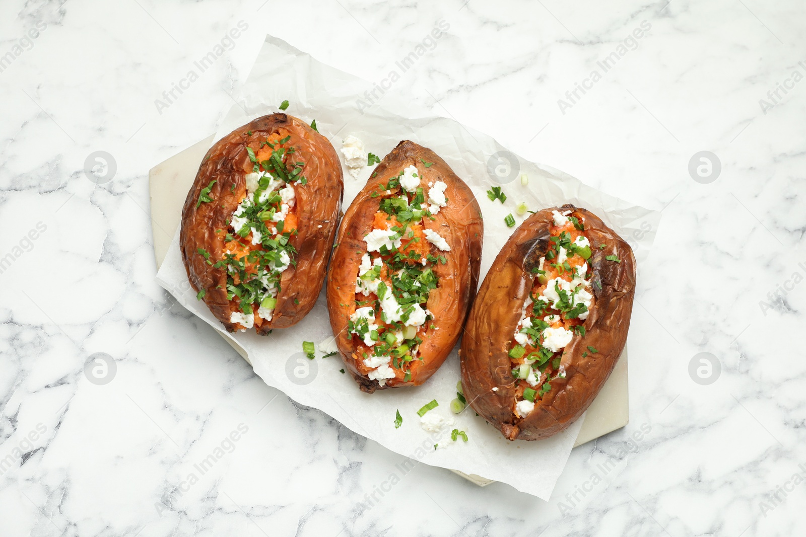 Photo of Tasty cooked sweet potatoes with feta cheese, green onion and parsley on white marble table, top view