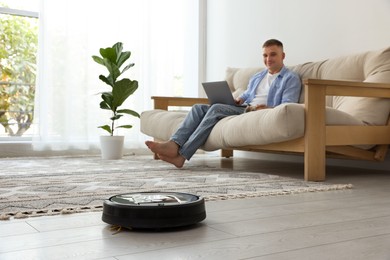 Photo of Young man using laptop while robotic vacuum cleaner vacuuming floor at home