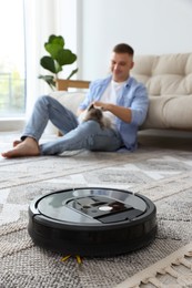 Photo of Young man with cute cat at home, focus on robotic vacuum cleaner