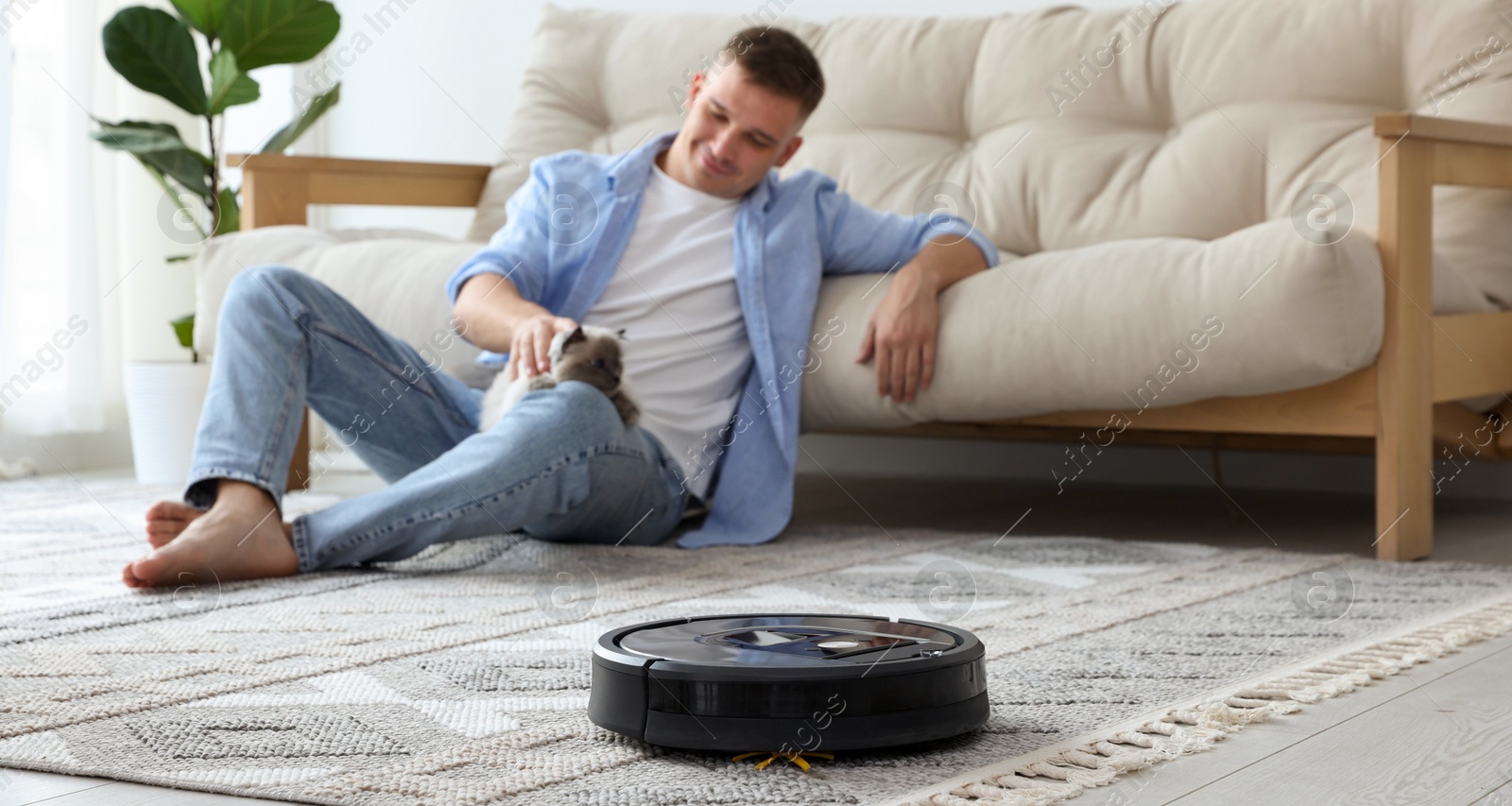 Photo of Young man with cute cat at home, focus on robotic vacuum cleaner