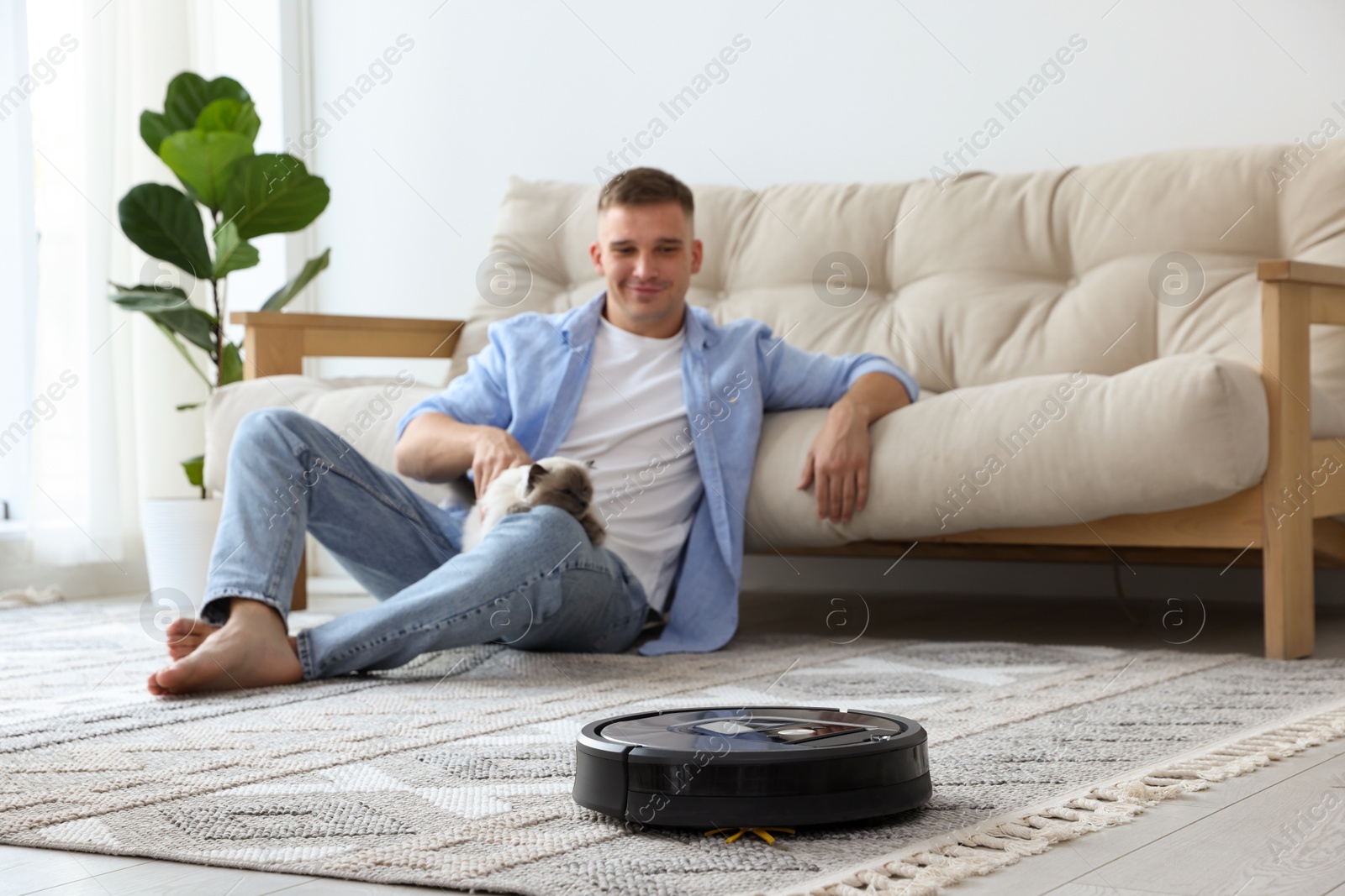 Photo of Young man with cute cat at home, focus on robotic vacuum cleaner