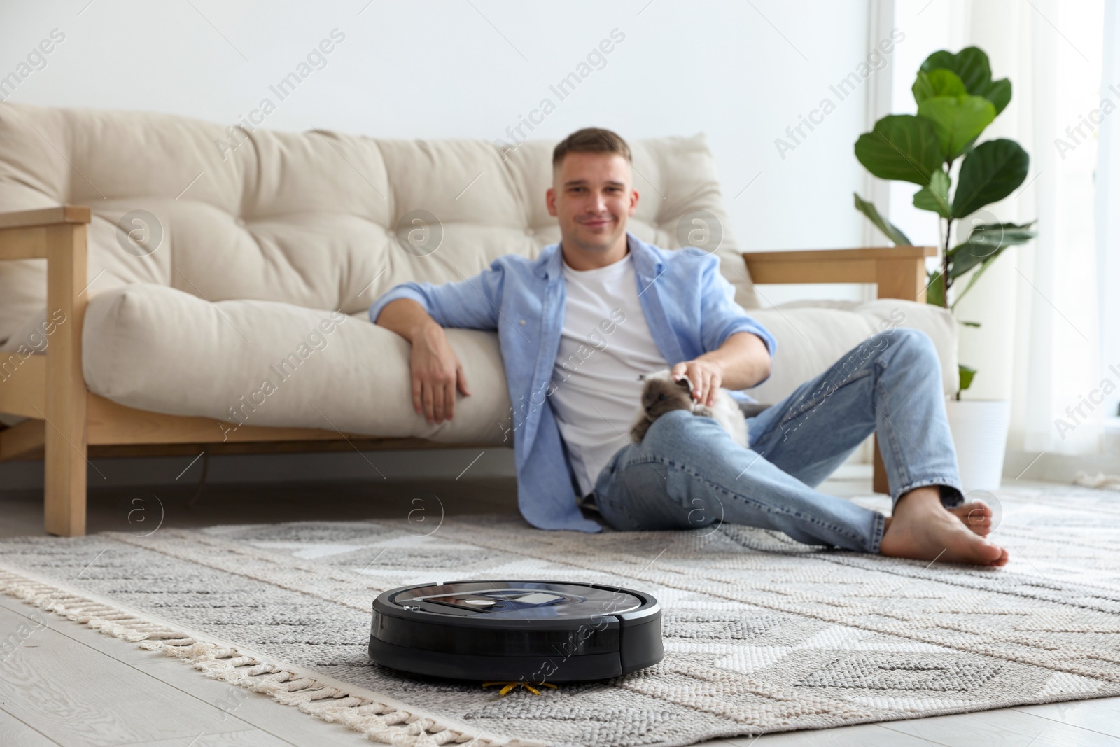 Photo of Young man with cute cat at home, focus on robotic vacuum cleaner