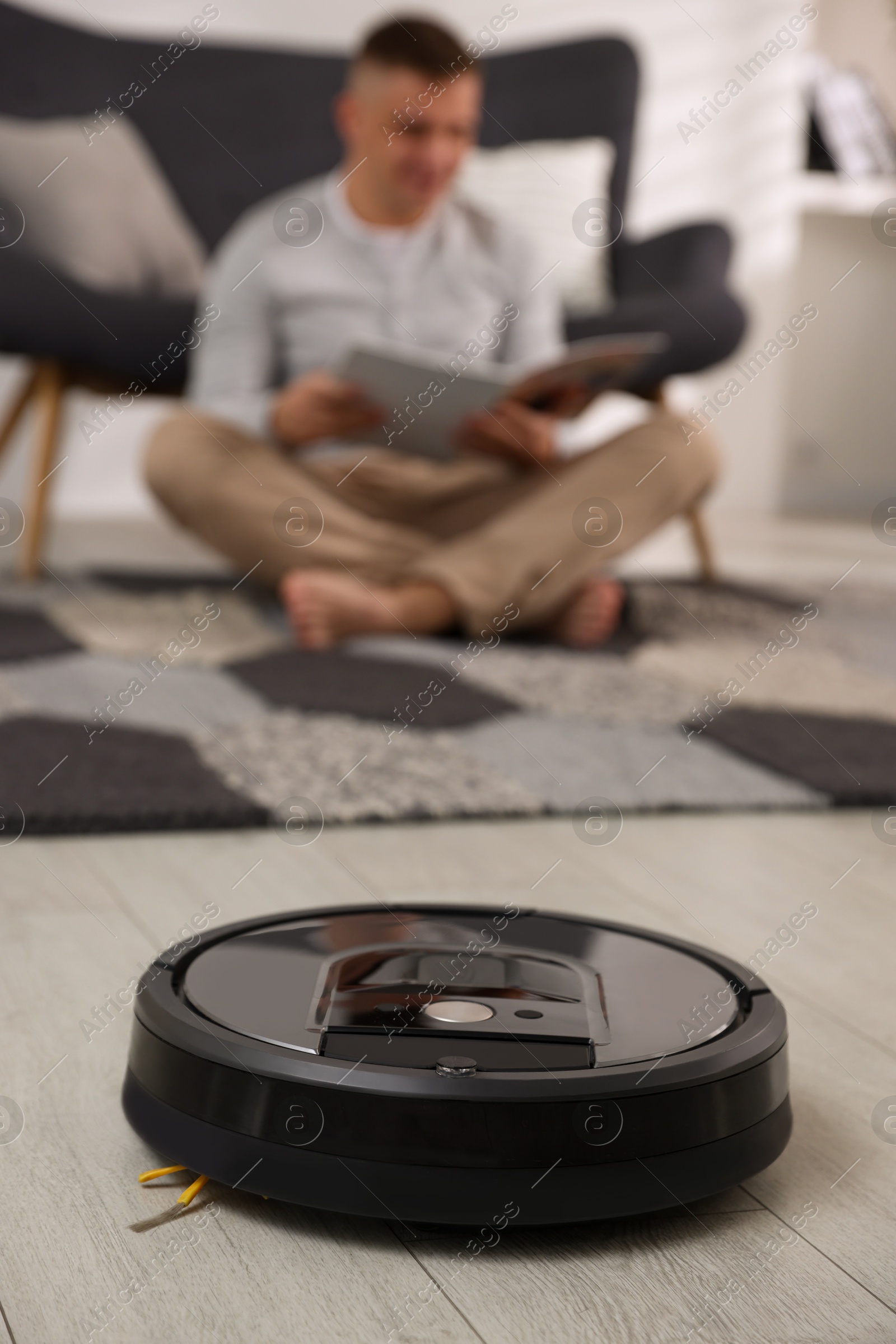 Photo of Young man reading magazine at home, focus on robotic vacuum cleaner