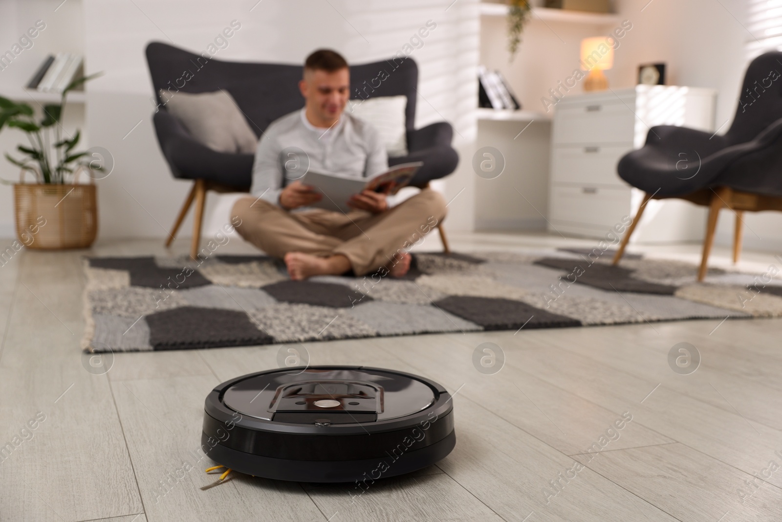 Photo of Young man reading magazine at home, focus on robotic vacuum cleaner