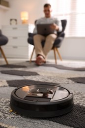 Photo of Young man using laptop at home, focus on robotic vacuum cleaner