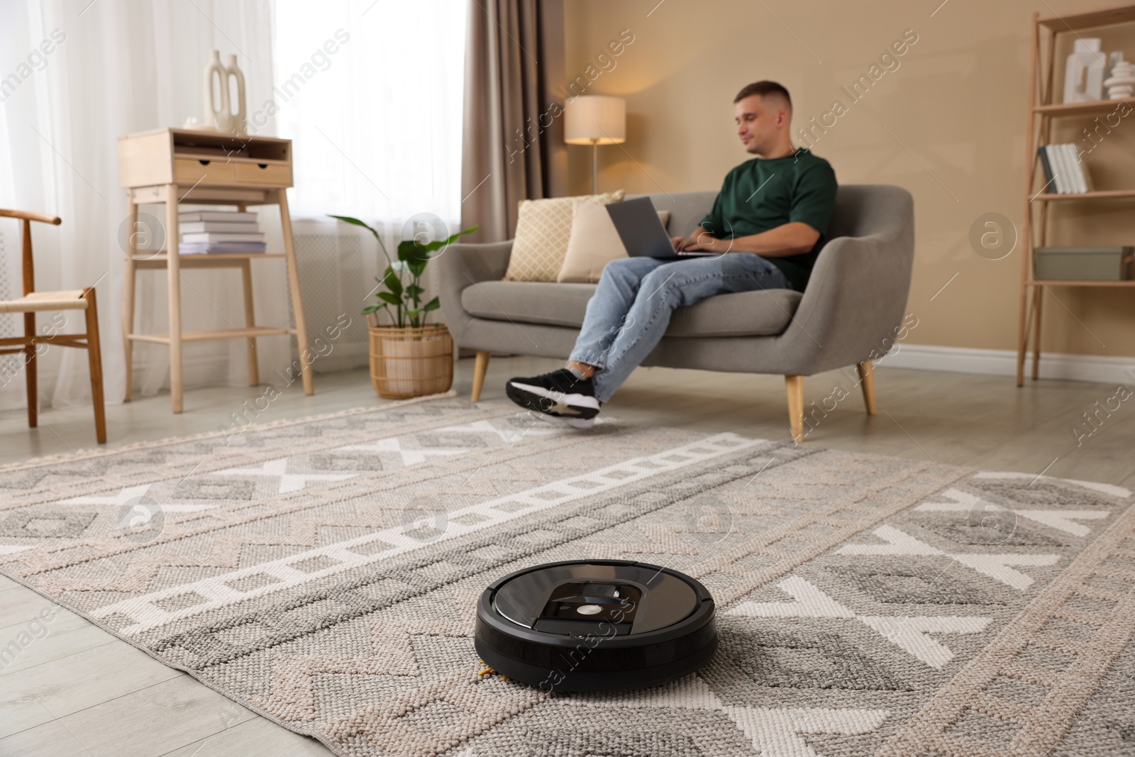 Photo of Young man using laptop at home, focus on robotic vacuum cleaner