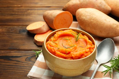 Photo of Tasty mashed sweet potato with rosemary in bowl, fresh vegetables and spoon on wooden table, closeup
