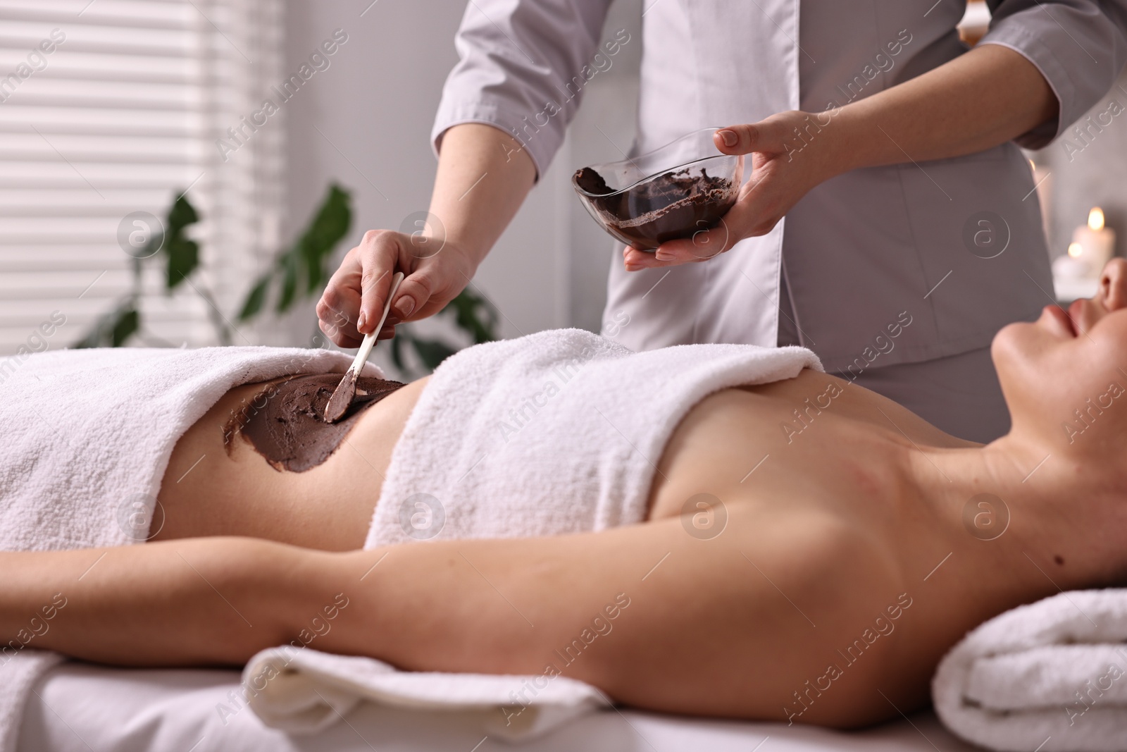 Photo of Chocolate body wrap. Spa worker applying mask onto woman's belly in salon, closeup