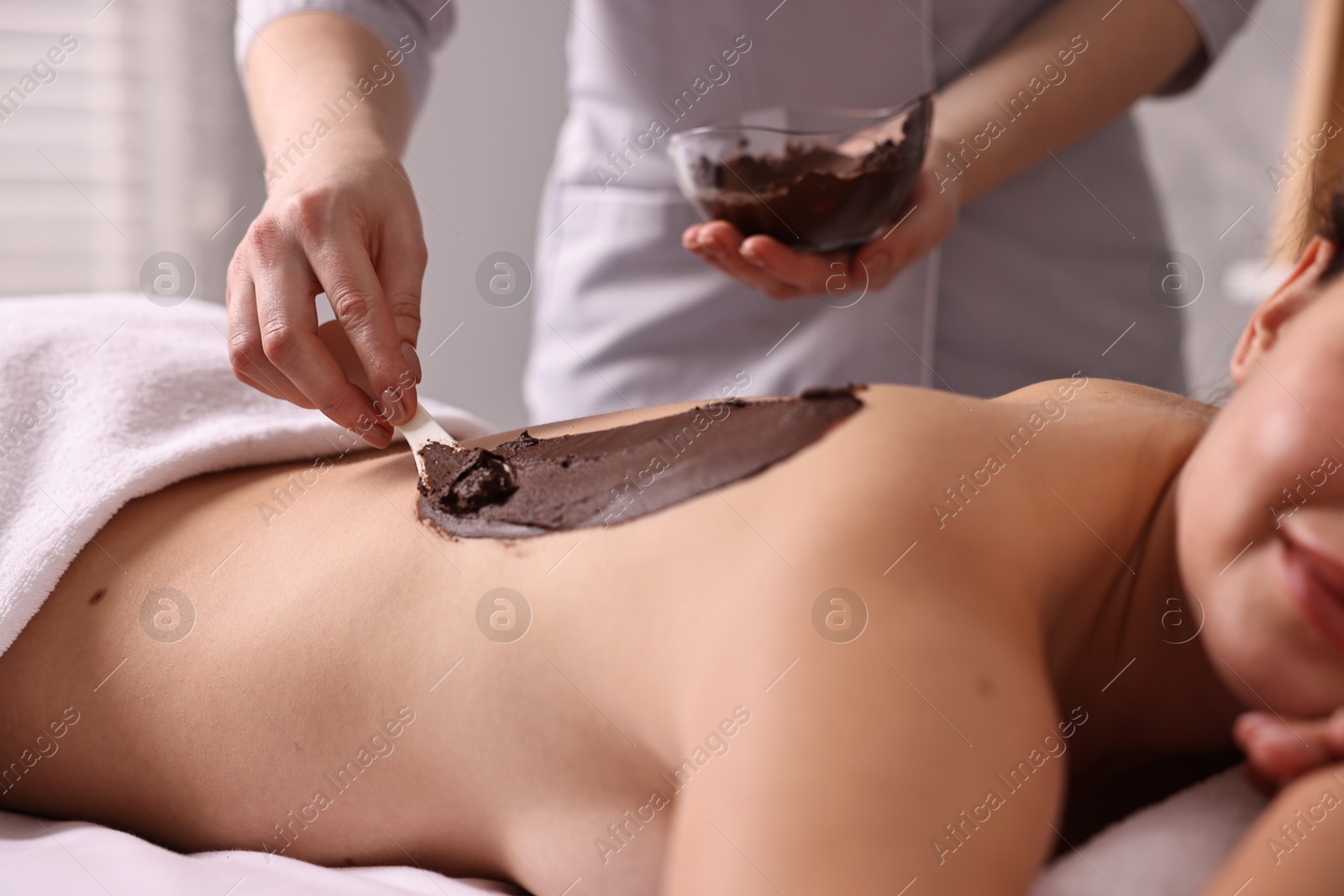 Photo of Chocolate body wrap. Spa worker applying mask onto woman's back in salon, closeup